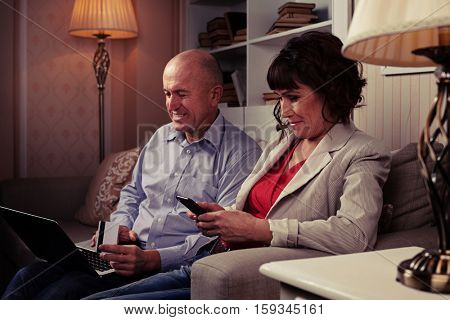 A mid side shot of smiling people sitting on the brown settee surrounded by lamps and booksettle. Man with laptop laughing and riant lady looking at her mobile