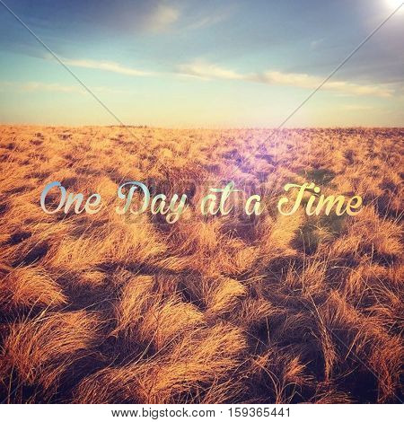 Inspirational quote.  One day at a time. Bright colorful field landscape with blue sky, clouds and orange colored grass in prairie field at sunset.    Colorful sky over horizon.  Instagram effects