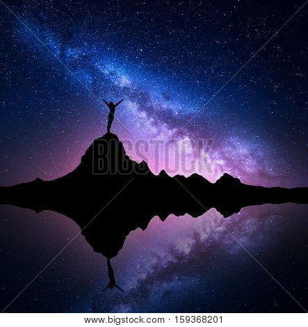 Milky Way. Night starry sky and silhouette of a standing woman with raised up arms on the mountain peak near the lake with reflection in water. Landscape with Milky Way and woman. Galaxy Universe