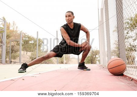 Photo of young african basketball player making sport exercises. Shooting at basket outdoor. Look aside.
