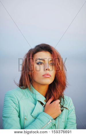 Portrait of beautiful brown-eyed girl brown-haired in turquoise blue leather jacket shivering from the cold wind on blurred background closeup