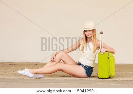 Travel adventure teenage journey concept. Woman wearing denim shorts white top and sun hat suitcase holding suitcase on wheels hitchhiking sitting and relaxing during trip