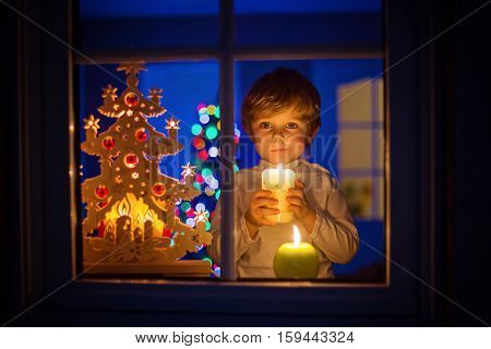 Beautiful little kid boy, standing by window at Christmas time and holding candle. Child waitng for Santa. With colorful lights from Christmas tree on background.