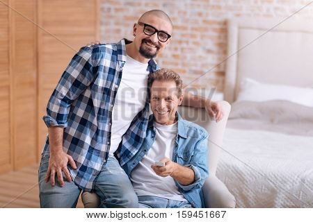 We happy together. Cheerful positive non-traditional couple sitting on the settee in the bedroom and watching television while hugging and expressing happiness