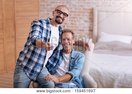 Switching the channel. Happy positive non-traditional couple sitting on the sofa in the bedroom and watching television while hugging and switching the channel