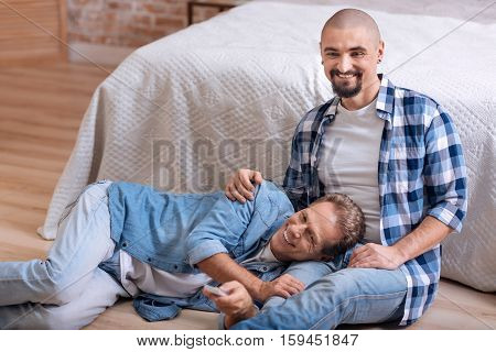 So nice to be close to you. Delighted positive smiling non-traditional couple sitting on the floor in the bedroom and watching television while hugging and switching the channel