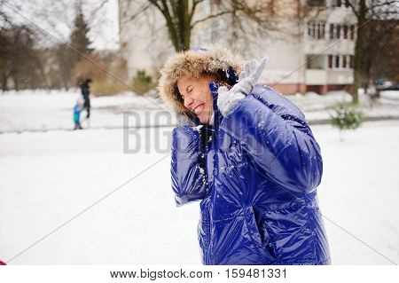 Woman turns aside from the snowballs flying to her. Someone fires at her snowballs. She pulls a hood over the face and cheerfully laughs. All ground is completely covered with fluffy snow.