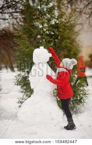 The girl of school age builds a snowman. On the ground there is a lot of snow. The girl in a red jacket and a gray knitted cap stands near a big snowman. She decorates the head of a snowman.