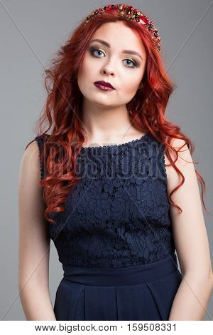 Beautiful red-haired fashion model posing in evening dress and in the diadem over dark background. female gestures of seduction. body language. touching her hair.