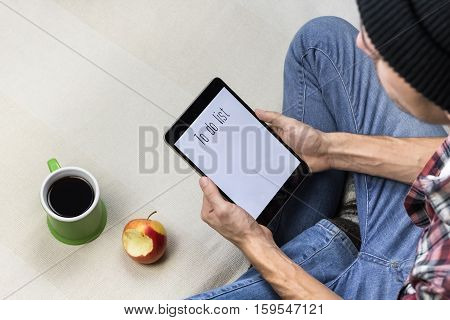 Making to do list. Male person in jeans and casual shirt works with tablet PC at home, having a coffee and apple snack