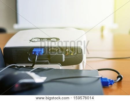 Closeup of projector for presentation connected to laptop with blank screen in front in sunny conference room