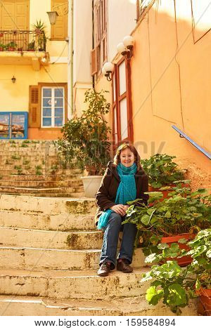 Smiling girl in blue scarf sitting on the stairs on a street of Greek town Nafplion on a sunny winter day