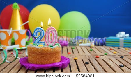 Birthday cake with candles on rustic wooden table with background of colorful balloons, gifts, plastic cups and candies with blue wall in the background. Focus is on cake