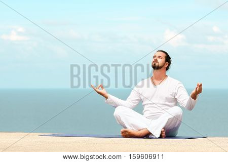 Caucasian man in white clothes meditating yoga on the sea shore pier