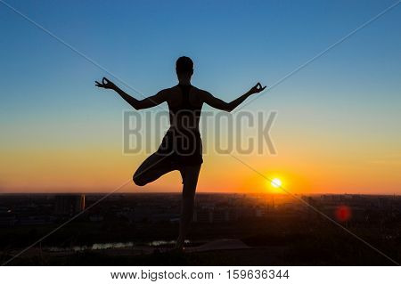 Silhouette of sporty woman practicing yoga in the park at sunset - half lotus tree pose. Sunset light, golden hour. Freedom, health and yoga concept