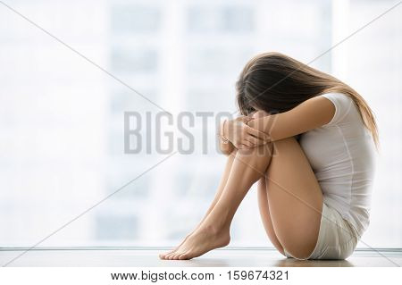 Young barefoot woman sitting at the floor embracing her knees, near full length window at home, her head down, bored, troubled with domestic violence, unwilling pregnancy, abortion, trolling