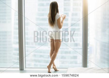 Full length portrait of young woman in casual home wear relaxing with cup of coffee in modern luxury apartment, with floor to ceiling window. Wakeup, dreaming, looking at rainy city scenery. Rear view