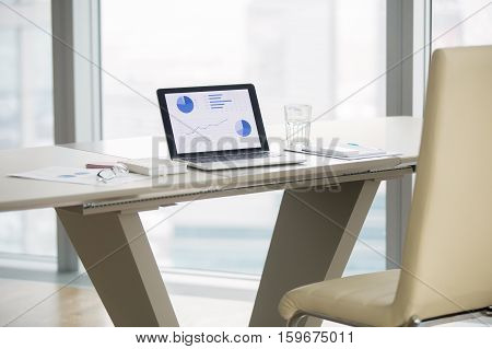 Laptop with graphs on screen on white modern office desk of business analyst, revealing business progress, seeking technology solutions, vacant place. Luxury office interior. Business success concept
