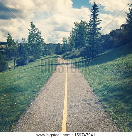 Park landscape paved trail and yellow line. Lush green grass and tall trees along trail. Blue sky and white clouds background.  Instagram effects.