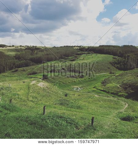 Field landscape in summer.  Lush dark green valley and hills with old wire fence and posts, bright lush green trees and grass. Bright blue sky, dark and white clouds background.