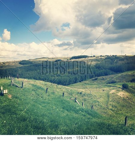 Field landscape in summer.  Lush dark green valley and hills with old wire fence and posts, bright lush green trees and grass. Bright blue sky, dark and white clouds background.