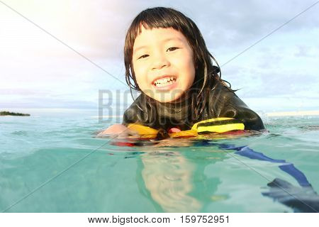 Happy little Asian girl playing with yellow life jacket in clear blue sea on hot summer day. Children wearing sun protection wetsuit relaxing in tropical sea.