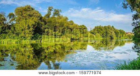 Summer warm evening. Panorama with river views from the footbridge. The village is in the background. Reflection of blue sky in the river water.