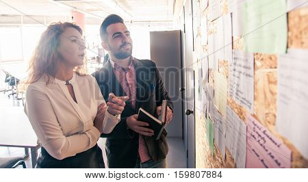 Businessman and businesswoman communicating while standing near notice board in office. People discussing following meetings in board room soom.