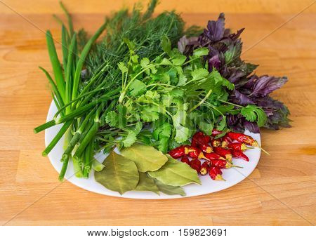 Close up of green organic herbs, basil, coriander, fennel, green onion, red chili peppers on white plate, rustic wooden background. Fresh herbs for Caucasian, Turkish, Mediterranean, Oriental cuisine.