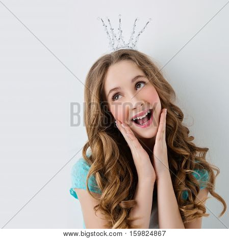 Happy teenage girl with long curly hair and crown on head looking like princess. Surprised expression. Studio portrait on light background. Copy space. Square composition.