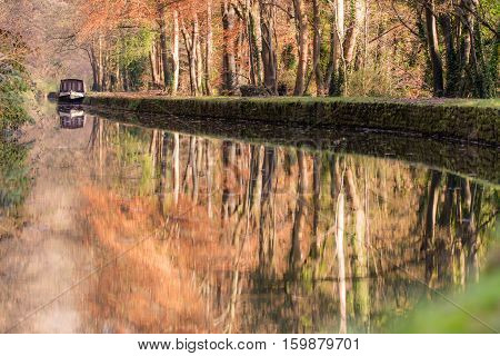 Narrow boat on Kennet and Avon Canal in autumn. Autumnal scene with orange leaves of beech trees reflected in still water of canal in English countryside