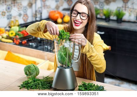 Young smiling woman making smoothie with fresh greens in the blender in kitchen at home. Healthy vegetarian smoothie for weight loss and detox