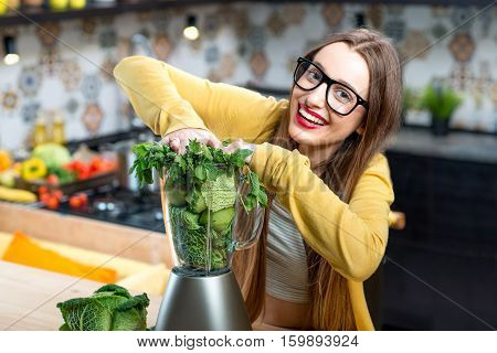 Young smiling woman pushing fresh greens into the blender in the kitchen at home. Healthy vegetarian smoothie for weight loss and detox