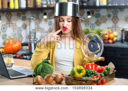 Unhappy woman with cooking pan on the head in the kitchen full of vegetables. Dissatisfaction with the process of cooking