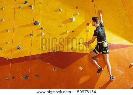 Extreme way of life. Fearless handsome young man climbing up the wall while spending time in climbing gym and using equipment.