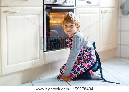 Cute funny blond kid boy baking muffins in domestic kitchen. Child having fun with helping, sitting near ofen and waiting for cupcakes.