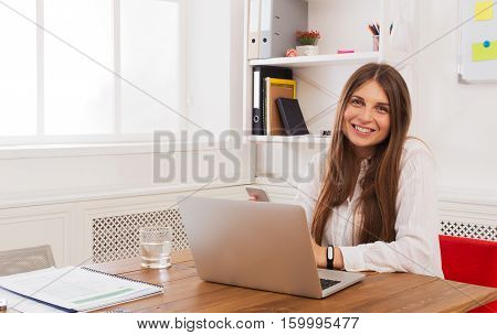 Beautiful young businesswoman sitting by wooden desk with laptop. Modern office worker in white room interior. Succesfull woman, female carreer concept.