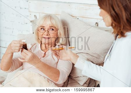 Improve your health. Pleasant sick elderly woman lying in bed and holding bottle with pills while professioanl doctor visiting her at home
