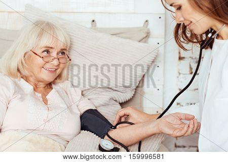 Professional attitude. Pleasant sick delighted elderly woman lying in bed and smiling while professioanl measuring her blood pressure at home