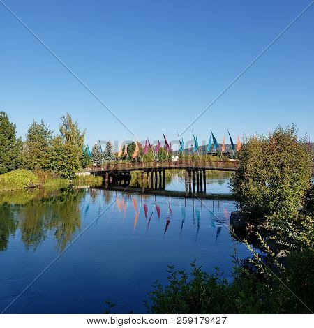 A Wooden Walking And Biking Bridge With Multi-colored Flags Crosses The Blue Waters Of The Deschutes