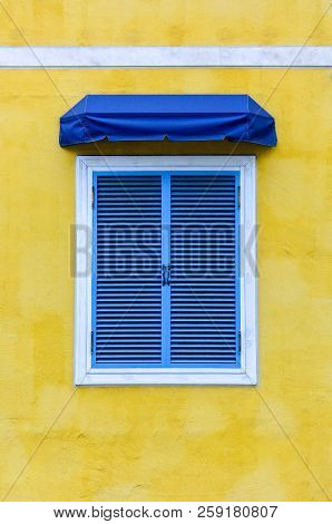 Closed Traditional Blue Greek Window With White Frame And Blue Fabric Awning On Yellow Cement Backgr
