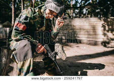 Smiling Young Female Paintballer In Camouflage Uniform Sitting With Paintball Gun Outdoors