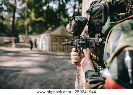 Selective Focus Of Paintball Player In Goggle Mask And Camouflage Aiming By Paintball Gun Outdoors
