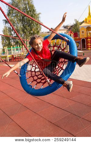 Happy Smiling Girl Swinging On A Swing With Closed Eyes And Arms Outstretched At Playground In Summe
