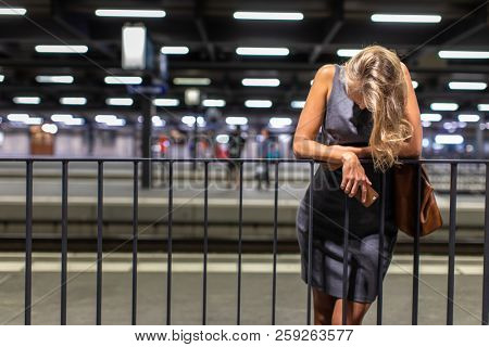 Exhausted overworked young female commuter waiting for her daily train in a modern train station, using her cellphone while waiting (color toned image)