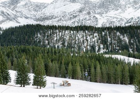 Curved Foothills And The Rocky Alps Mountains Covered By Snow And Green Fir Trees, A Cableway System