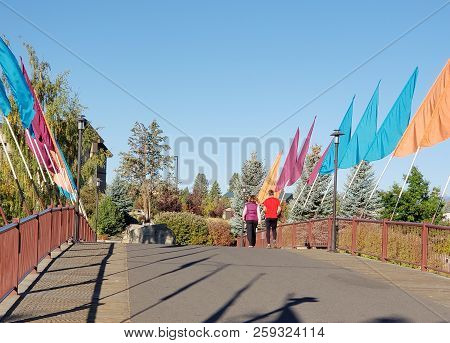 A Couple Walks Across A Wooden Walking And Biking Bridge With Multi-colored Flags That Crosses The D
