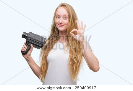 Blonde woman teenager filming holding super 8 video camera doing ok sign with fingers, excellent symbol