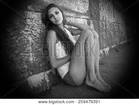 Portrait Of Sexy  Slender Young Woman In White T-shirt, Denim Shorts, Sitting Near Wall In An Erotic