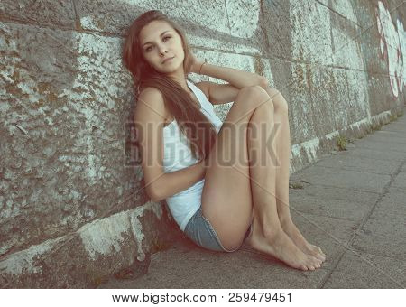 Portrait Of Sexy  Slender Young Woman In White T-shirt, Denim Shorts, Sitting Near Wall In An Erotic